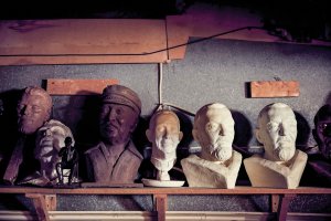 A slew of sculpted faces on a wooden shelf, along with a statuette of man sitting in a chair