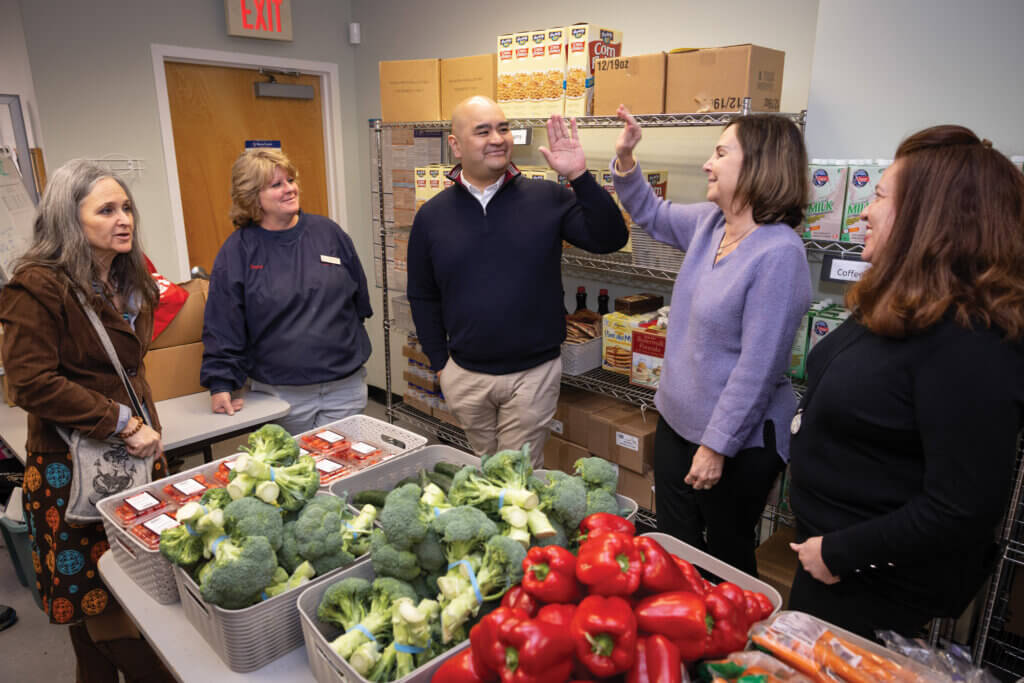 Five individuals standing in a food pantry, with fresh vegetables on a table in front of them and nonperishables on shelves behind them.