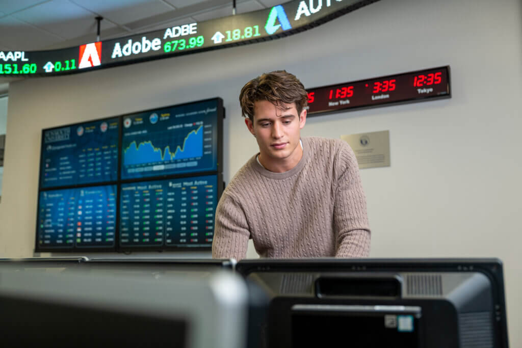 a student looks at a terminal in the financial markets lab, with stock tickers on screens behind him