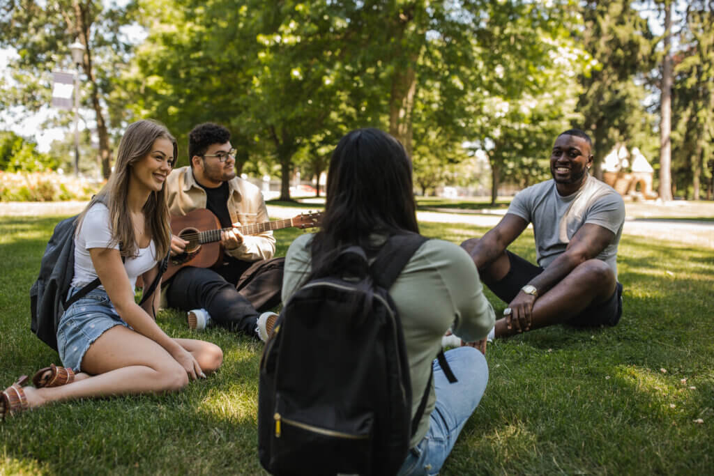 4 students, 1 with a guitar, sit in the grass on Shadow Lawn