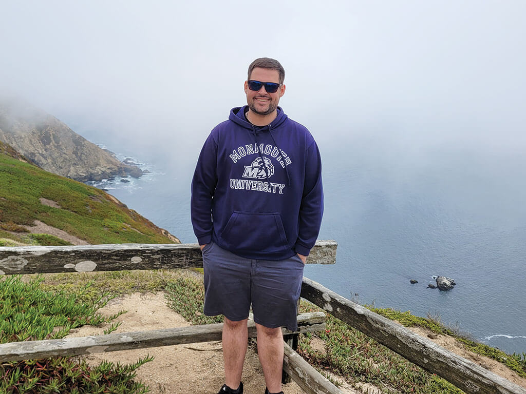 Matt Francis stands on a foggy coastal overlook wearing a Monmouth University sweatshirt and sunglasses.
