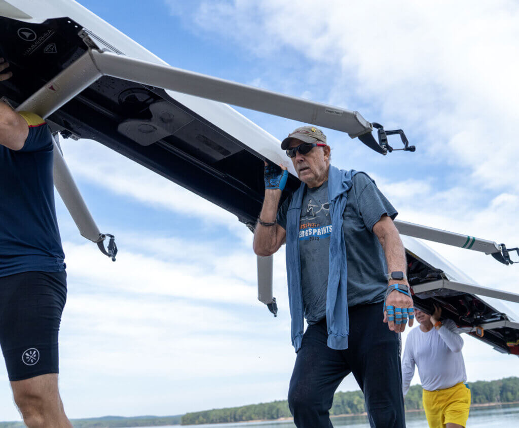 Ken Boyle carries a rowing shell with teammates near the water at Jordan Lake.