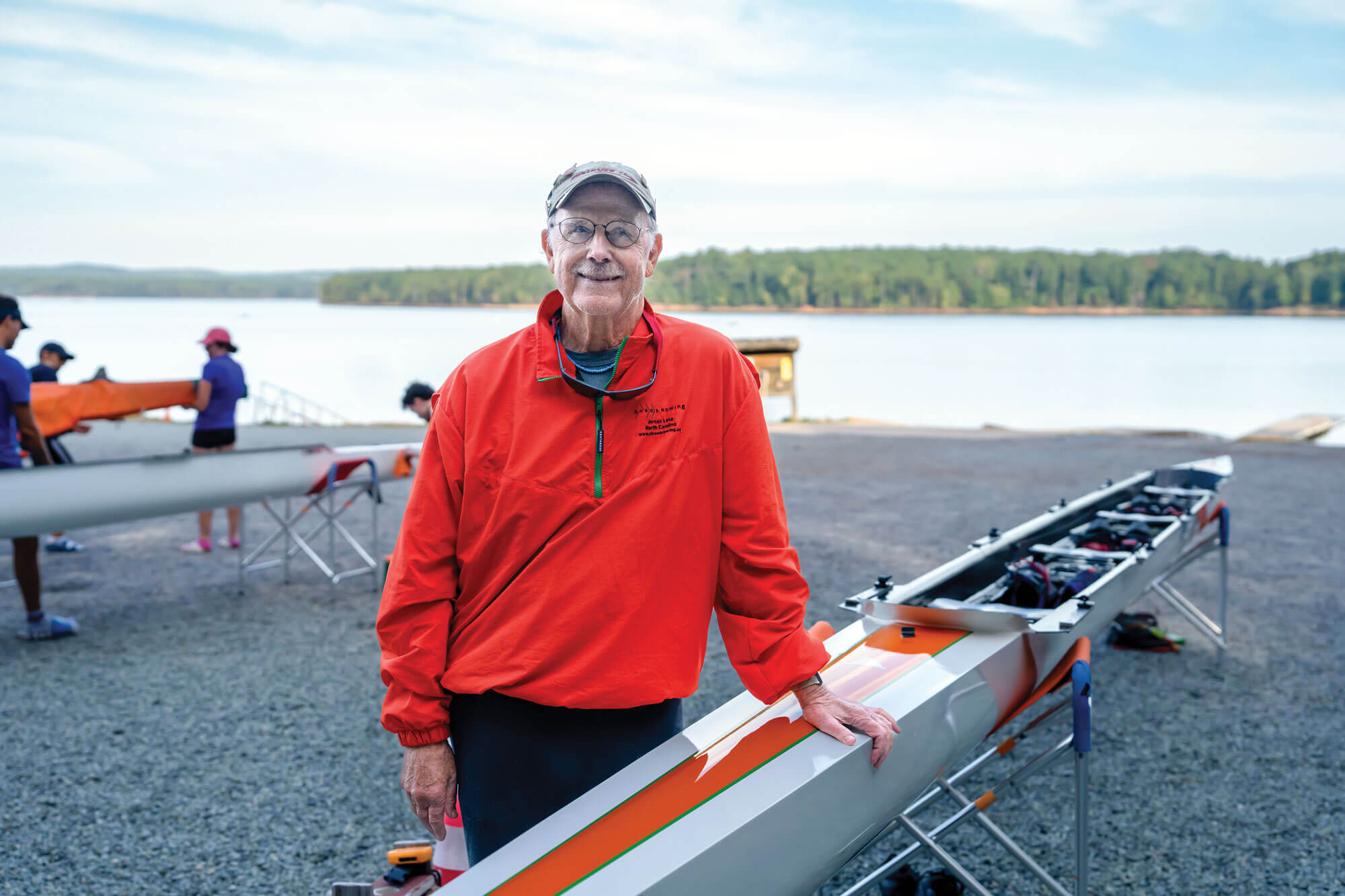 Ken Boyle standing beside a rowing shell with a lake and other rowers in the background