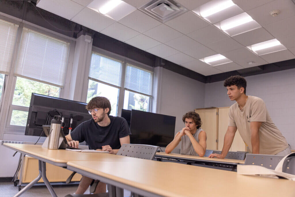 Three Monmouth University students collaborate in a classroom, looking at a laptop and discussing data