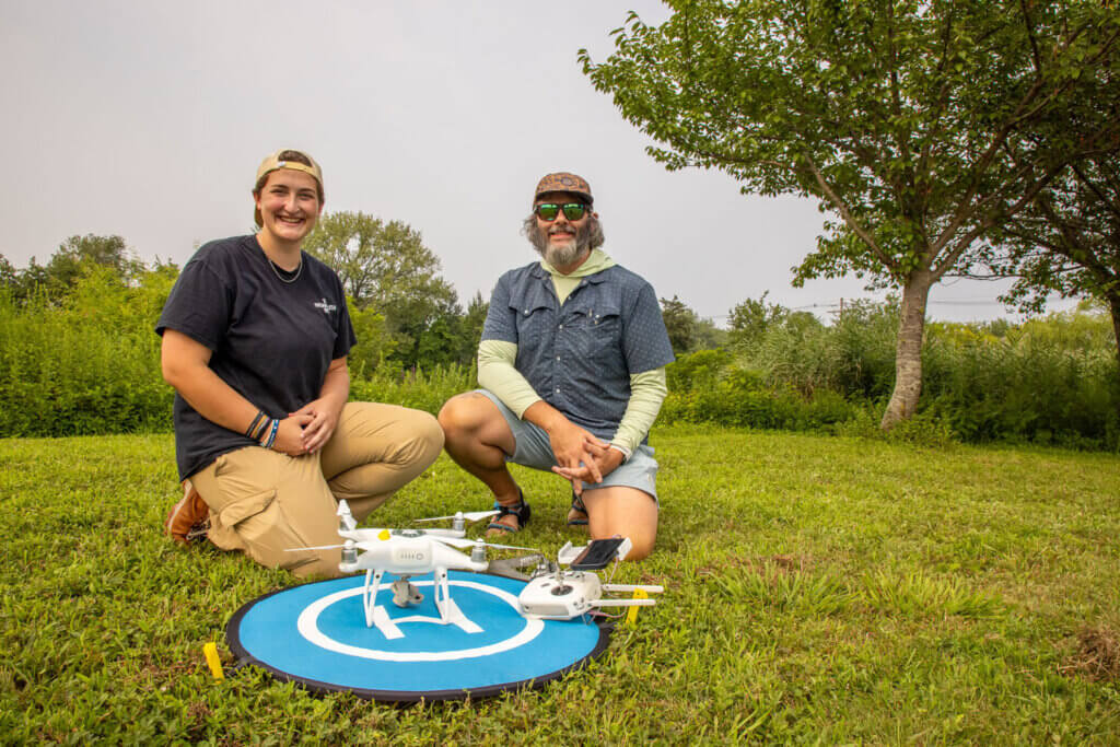Vicky O’Malley and Sean Sterrett kneel on a grassy field beside a small white drone positioned on a blue landing pad. Trees and shrubs line the background