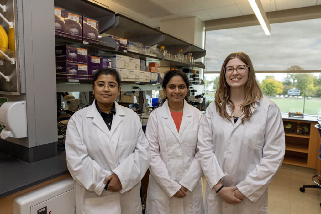 Saheli Sengupta, Aiswarya Raghavaraju, and Sarah Henry wearing lab coats stand together in a campus biology lab