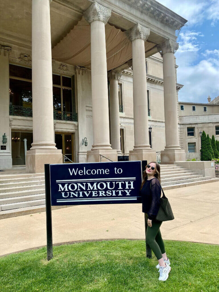 Megan Van Tine smiling beside a “Welcome to Monmouth University” sign in front of the Great Hall