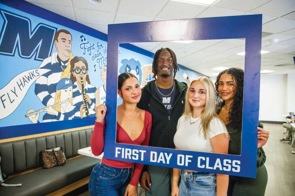 A photo of four first year students standing in the Rebecca Stafford Student Center holding up a sign that says "First Day of Class" with a big square cutout in the middle so that you can see their faces.
