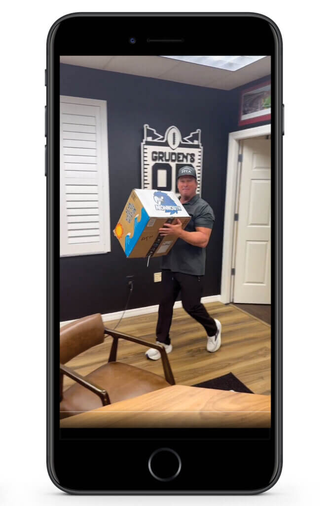 An image of an iPhone showing a screenshot of coach Jon Gruden walking with an unopened box of Monmouth Football swag. 