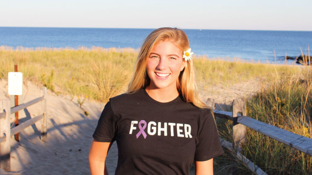 Alayna pictured on a beach with a flower in her hair and a shirt that says Fighter during her first battle with cancer when she was 17. 