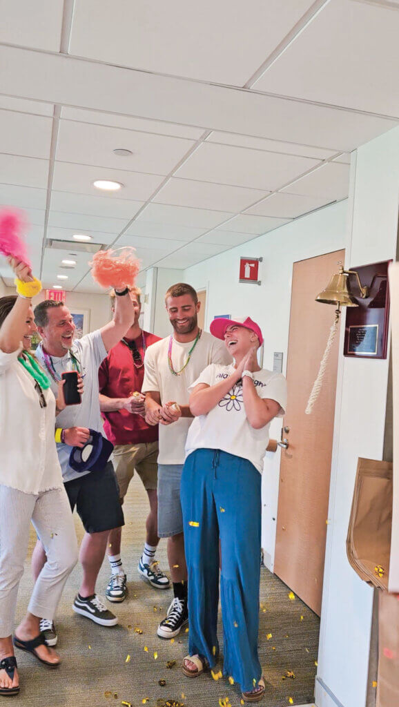 Alayna, learning she was cancer-free—for a second time—in 2024. She's standing surrounded by family and friends after ringing the bell in the hospital. 