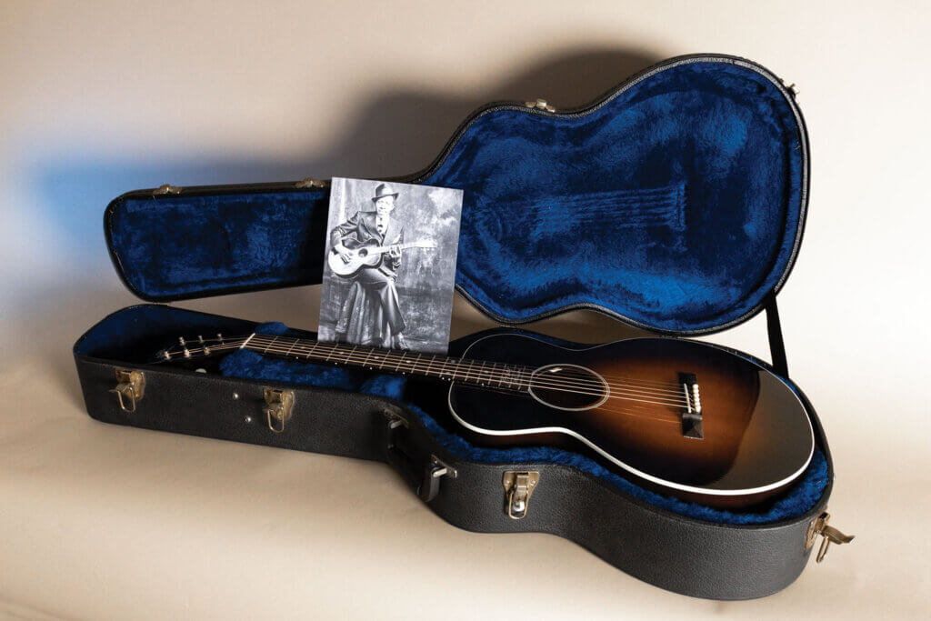 Acoustic guitar displayed in its open case alongside a historic photograph of blues musician Robert Johnson.