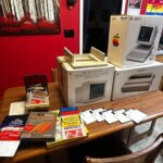 An Apple IIc computer system with monitor, printer, software, manuals, and floppy disks displayed on a table.