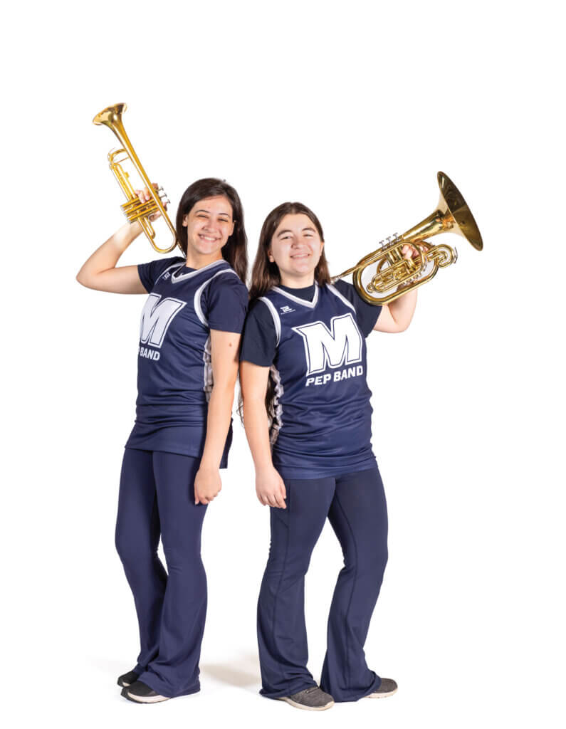 Daphne Valenzuela and Camryn Moschitta stand back-to-back in uniform, smiling while holding brass instruments