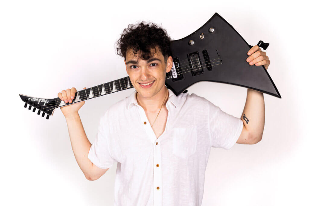 Leo Hernandez smiles while holding a black electric guitar over his shoulders