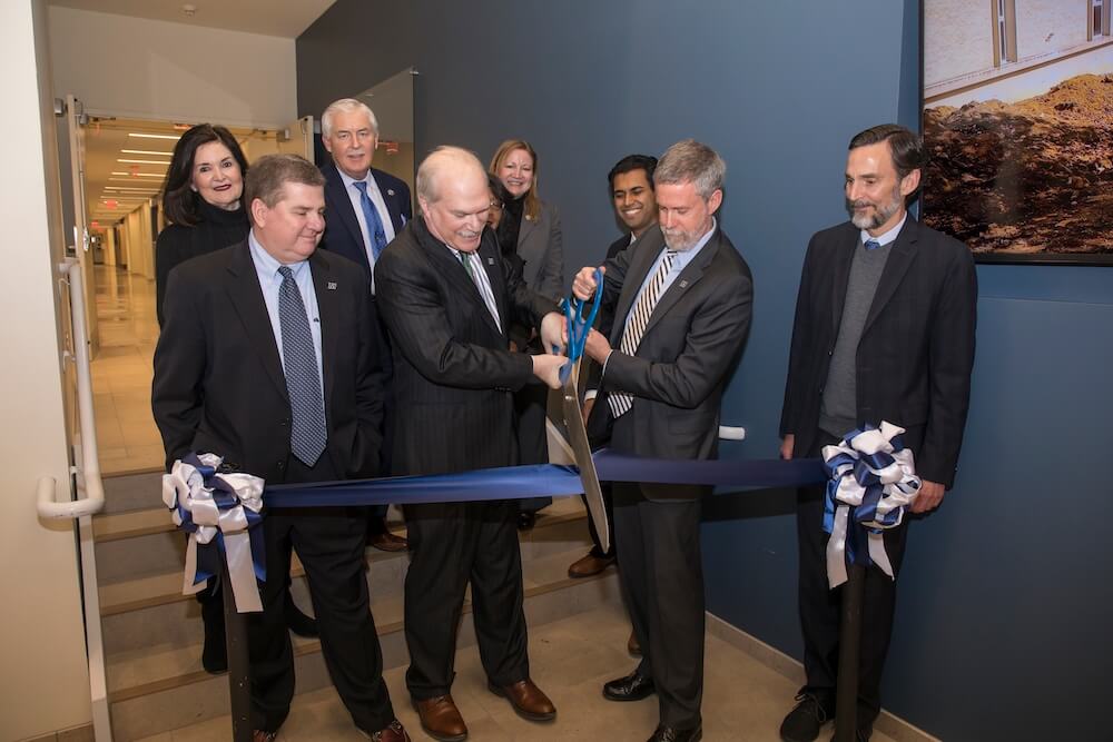 Photo of ribbon cutting; Pictured are School of Science Dean Steven Bachrach and University President Grey Dimenna, Board of Trustees Chair Michael Plodwick (l) and former Board Chair Henry Mercer, III, flank the pair. They are surrounded by (l-r) West Long Branch Mayor Janet Tucci, State Assemblyperson Eric Houghtaling, Superintendent of Higher Education Rochelle Hendricks (partially obscured), State Assemblyperson Joann Downey, and State Senator Vin Gopal.