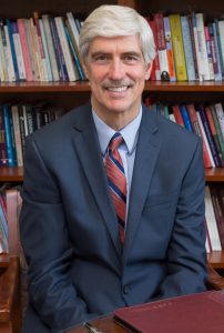 A photo of John E. Henning, sitting in front of a full bookcase.