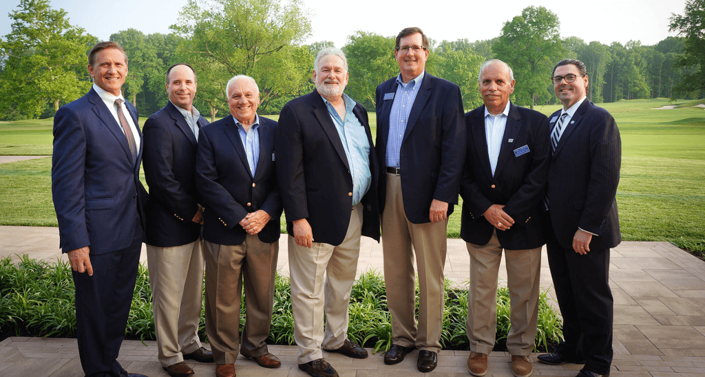 A group of seven men standing next to each other posing for the photo. There is a golf course in the background. 