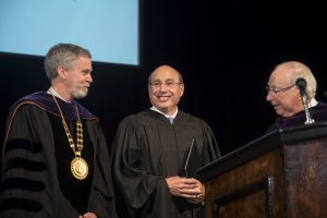 Three men behind a podium, the one in the center is smiling, all are wearing graduation gowns.