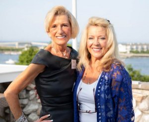 A photo of Carol Stillwell and Valerie Montecalvo posing for a photo standing side by side with a small harbor in the background. 