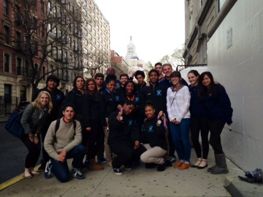 A group of people posing together on a sidewalk in a city, two of the people are holding up medals, 