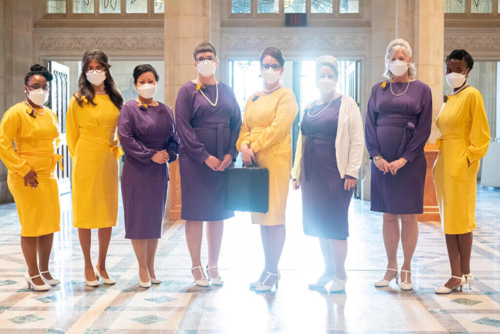 Eight 1960s-styled secretaries stand in a line, wearing yellow and purple dresses. One at the center holds a typewriter.