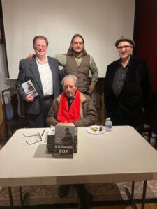 Three men standing behind seated man posing with book he authored