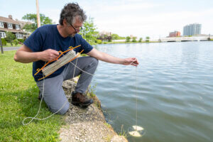 Prof. Adolf squats near edge of lake to perform water quality testing by dipping equipment on a string into water.