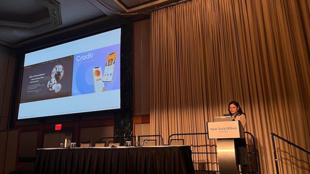 Female standing at podium marked "New York Hilton" in right side of image. On left side is screen showing graphic images representing mobile app "Cradlr".  Background is a pleated curtain.