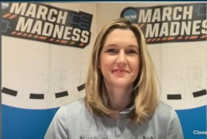 Women's Head Basketball Coach Ginny Boggess stands before March Madness signs