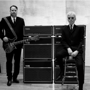 Two men in black suits stand in front of speakers. Man on left is holding a guitar.