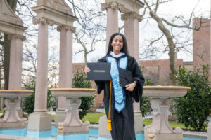 Monmouth University graduate in cap and. gown holding diploma in front of Erlanger Garden fountains after the commencement ceremony
