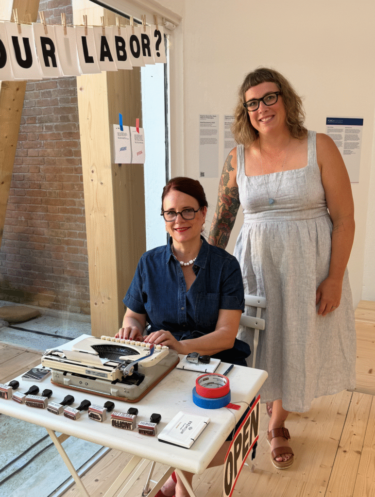 A woman sits at a typewriter as another woman stands behind her, both smiling at the camera.
