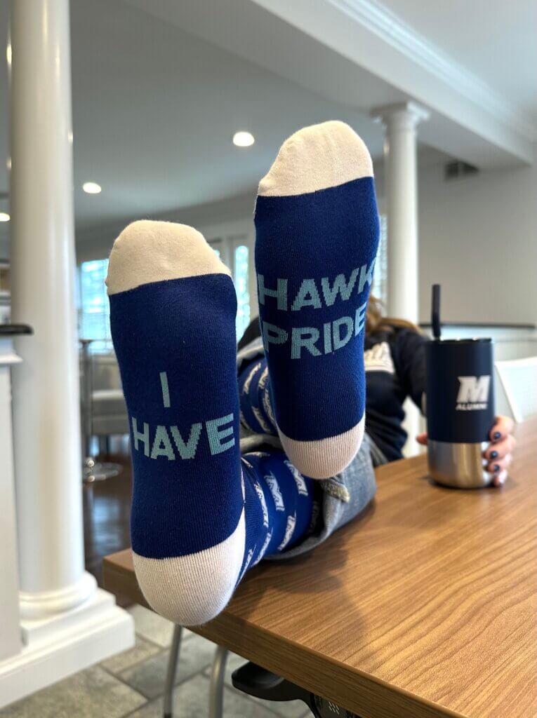 Image of a person wearing Giving Days socks. Feet crossed up on a table. The socks are navy blue with white spirit M's and text on the soles that reads "I HAVE HAWK PRIDE" in all caps.