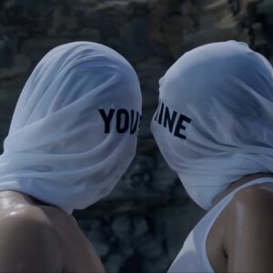 Two figures wearing white hoods over their heads stand in close proximity to one another.