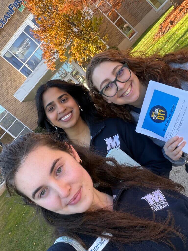 Three female OTD students on the campus of Monmouth University. One is holding a brochure that says, "Why OT?"