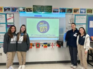 Four female students wearing Monmouth University clothing stand in front of a presentation about choosing Occupational Therapy