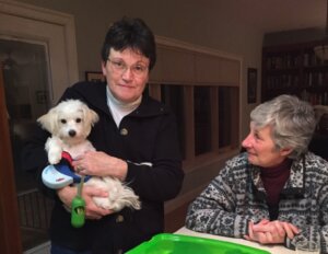 Two women at table. One is sitting, one is standing and holding a small white dog