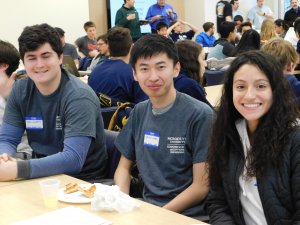 3 students (2 male and 1 female) sitting beside one another, smiling and eating lunch amidst a room of male and female program participants and their advisers