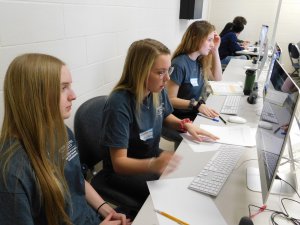 3 female students sitting along side one another with pens and paper on their desk in front of their computers