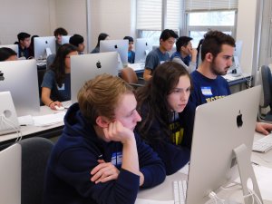 Room full of male and female students working on their programs at the competition with three students, two males and one female in the middle of the two, in the front of the photo