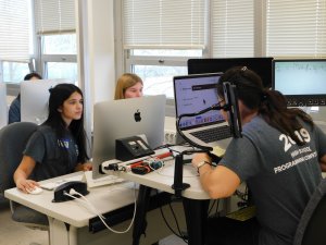 Three female students, 2 facing the camera and 1 facing her computer at the competition