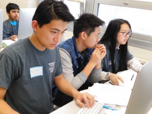 2 male students and 1 female students focused and working on writing programs at the competition with pens and papers on their desks