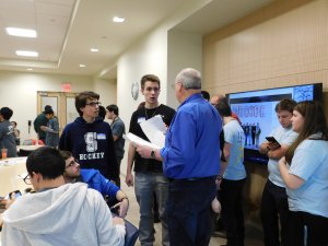 2 male students talking with their high school programming teacher, as other contestants and MU students stand nearby.