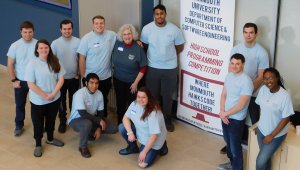 a group of 7 males and 3 female Monmouth University student volunteers with professor Kretsch. The group is posing in front of a banner for the 2019 High School Programming Competition