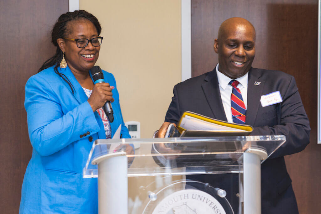 Two professionals at a podium, the woman on the left speaking in the microphone while the man to the right smiles