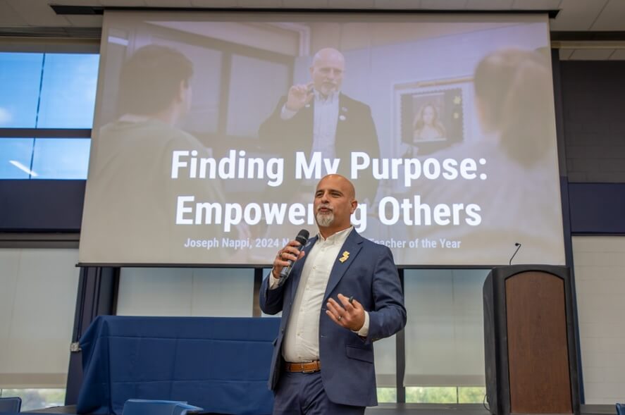 Man in suit speaking on a stage, in front of a screen that reads "Finding My Purpose: Empowering Others. Joseph Nappy, 2024 New Jersey Teacher of the Year"