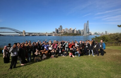 A large group of people standing in front of a body of water, with a bridge and city skyline behind them.