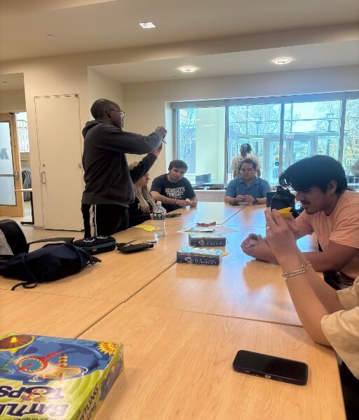 A man standing around a huge table, three man and two women sitting around the table, making paper origami.