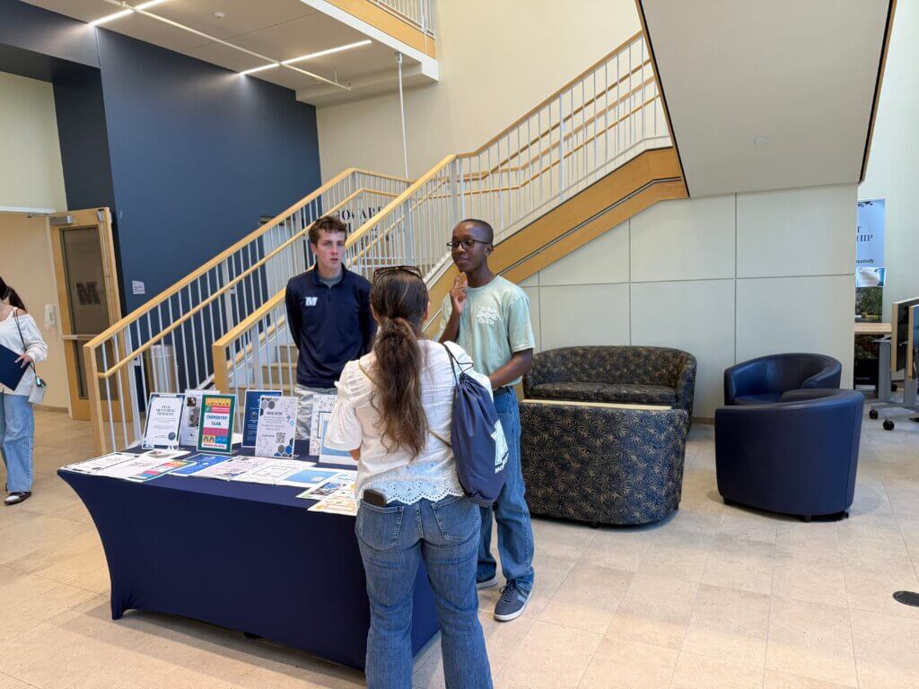 Two men stand in front of a table, talking to a women.